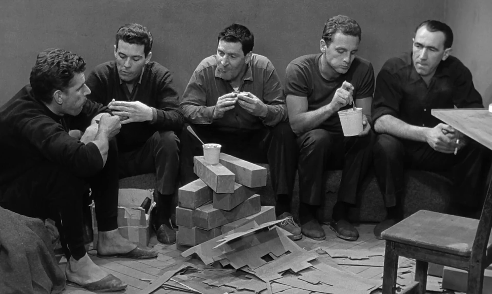 The five main characters of Hole eating in their prison cell in middle of their prison work which involves assembling cardboard boxes.